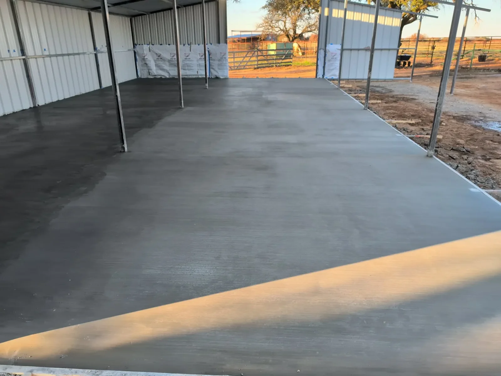Finished gray concrete slab with metal frame structure and corrugated metal roofing in agricultural setting