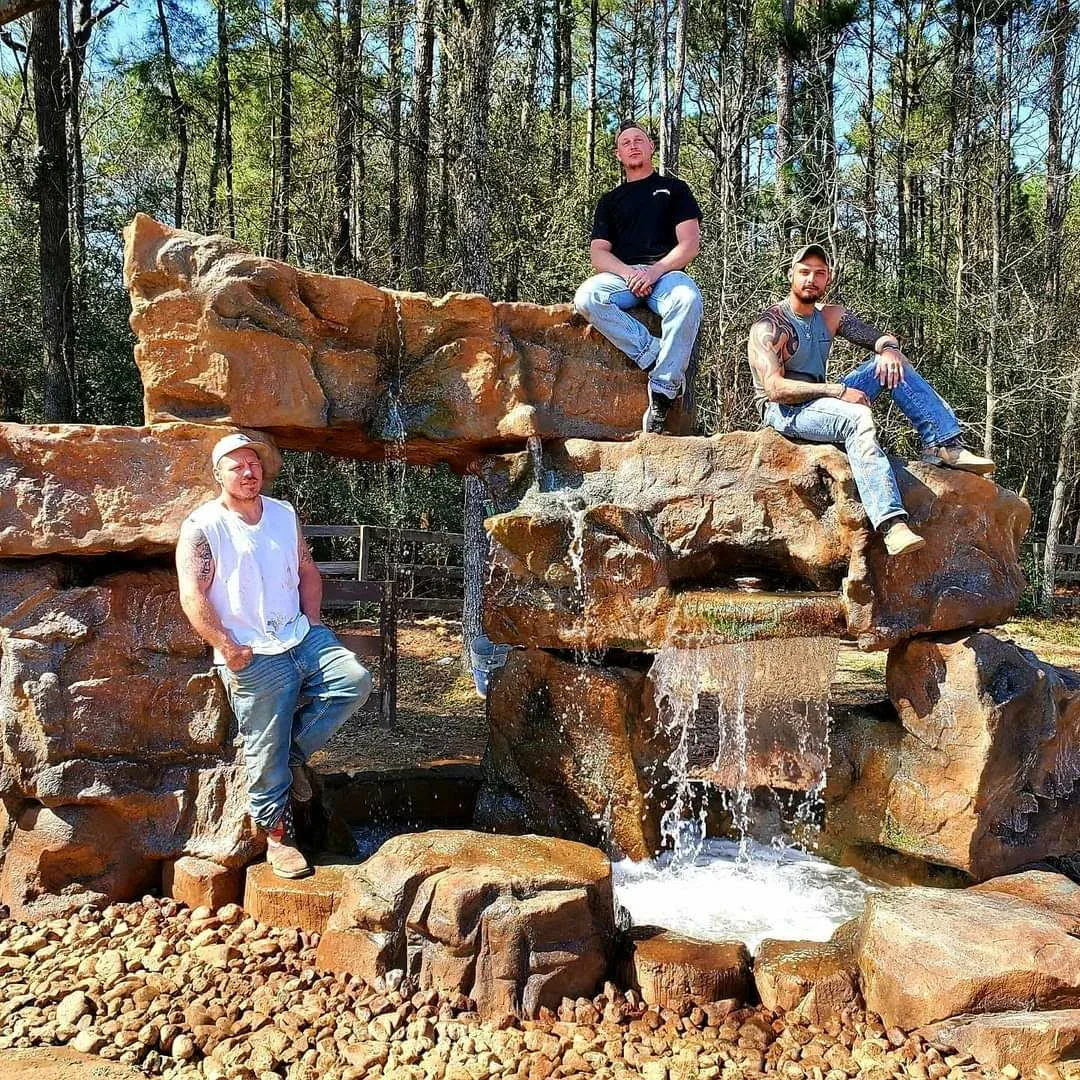 Stacked natural stone water feature with cascading water, river rock base, and concrete integration in wooded setting